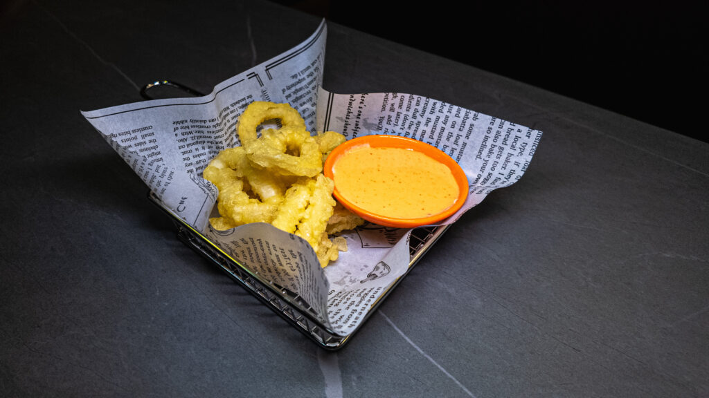 Golden fried onion rings served with orange cheese dipping sauce on newspaper in metal basket at restaurant