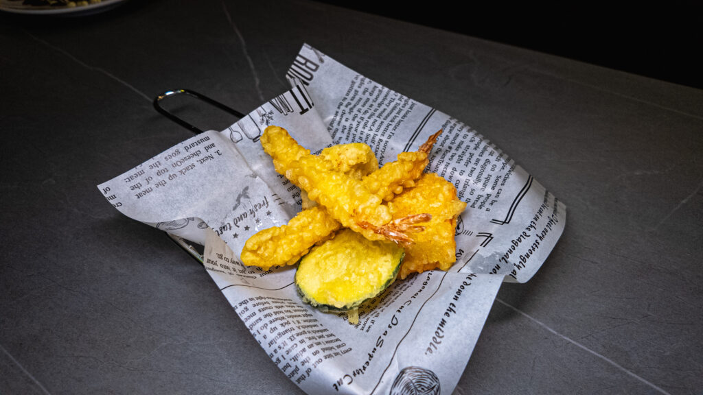 Golden fried tempura vegetables served on newspaper wrapping at restaurant on dark tile surface