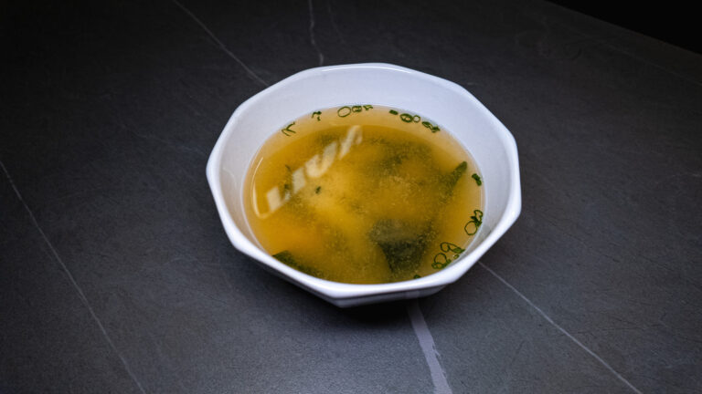 Bowl of clear broth soup with green herbs served in white ceramic bowl on dark tile surface at restaurant