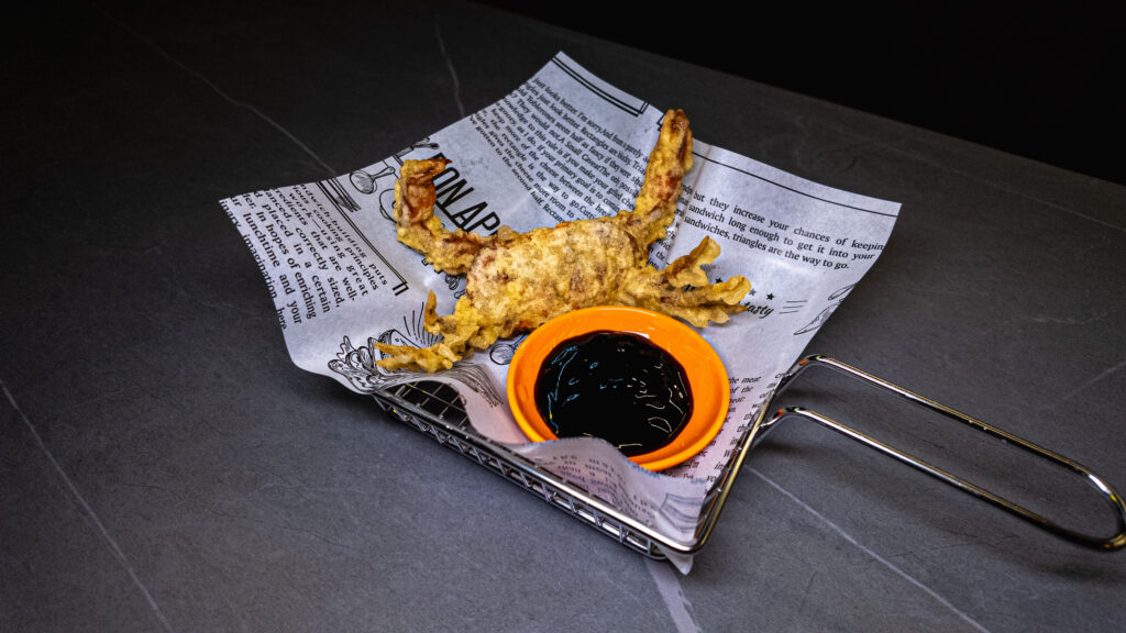 Deep-fried soft shell crab with dark dipping sauce served on newspaper in metal basket at restaurant