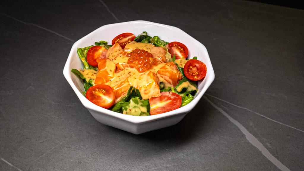 Fresh salmon salad with cherry tomatoes, mixed greens, and orange caviar in white octagonal bowl on dark slate surface