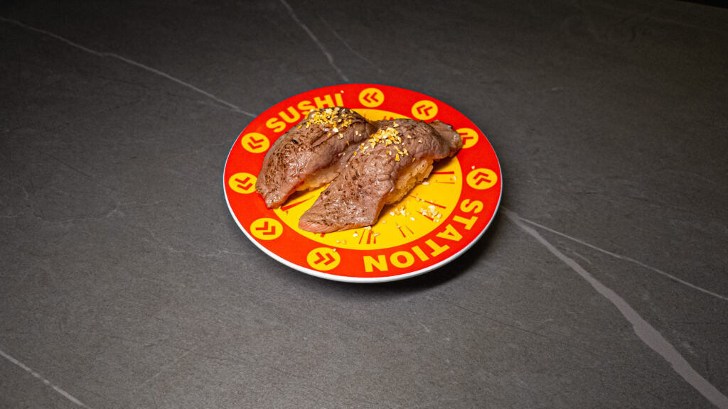 Three pieces of grilled beef nigiri sushi with seasoning on red conveyor belt plate at Japanese restaurant