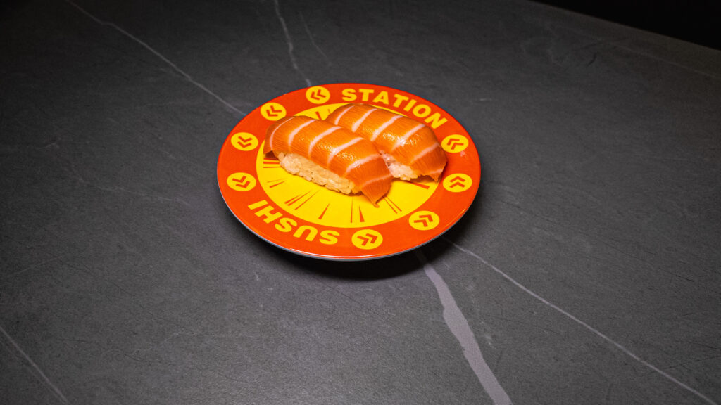 Salmon sushi nigiri served on orange Sushi Station branded plate at restaurant on dark slate surface