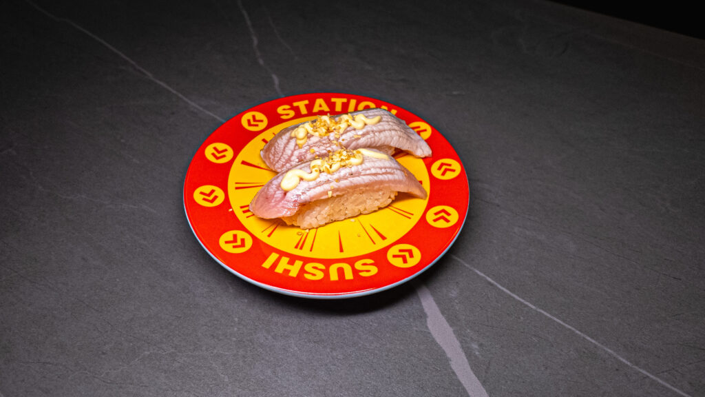 Two pieces of seared fish sushi with minced garlic on red conveyor belt sushi plate at restaurant