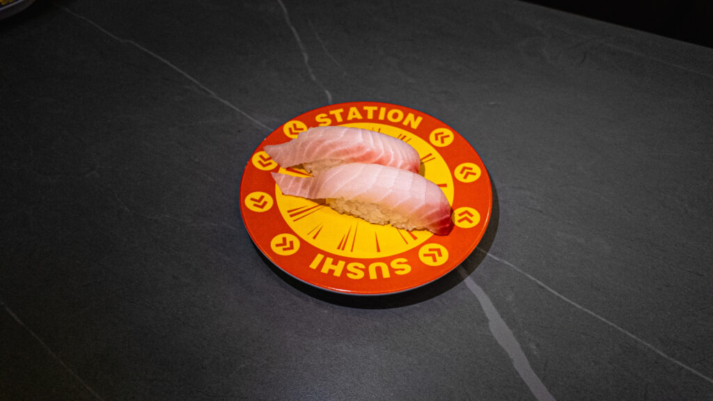 Two pieces of tuna sushi on orange Sushi Station branded plate on dark tiled floor at restaurant