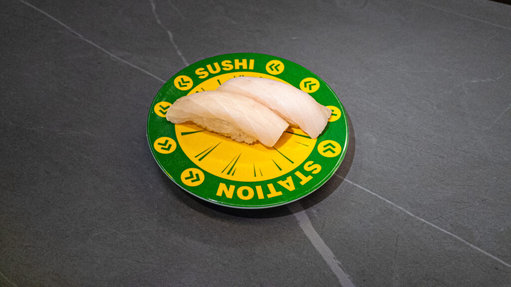 Two pieces of white fish sushi on green and yellow conveyor belt sushi station plate at restaurant
