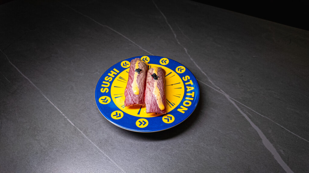 Two pieces of grilled meat with yellow sauce on blue SUSHI STATION branded plate on dark tile surface