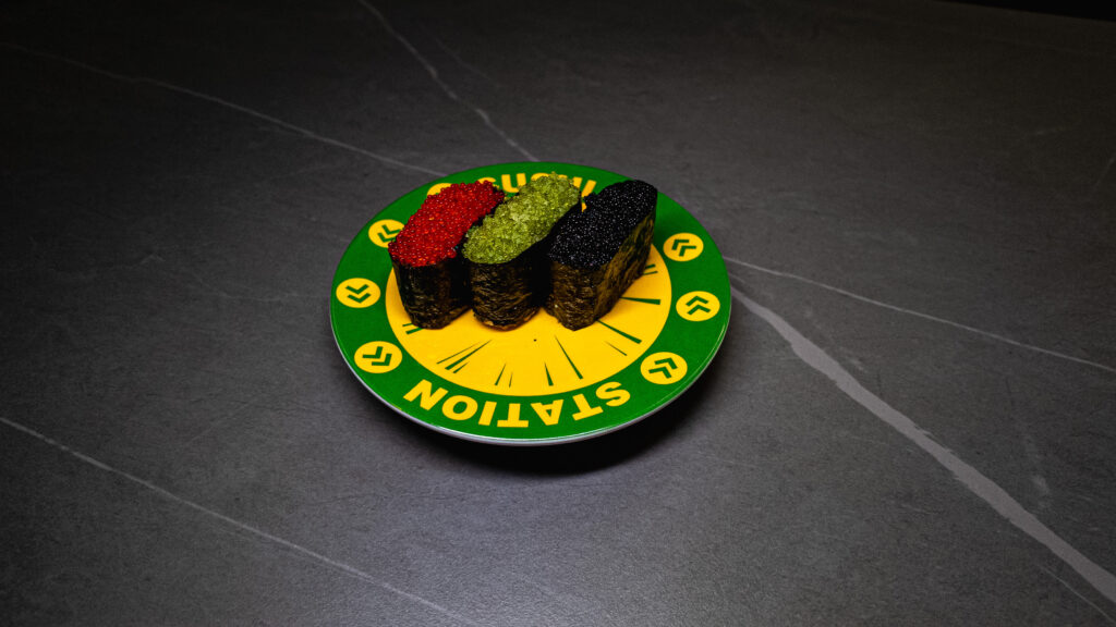 Three sushi pieces with colorful toppings on green conveyor belt sushi restaurant plate against dark marble surface