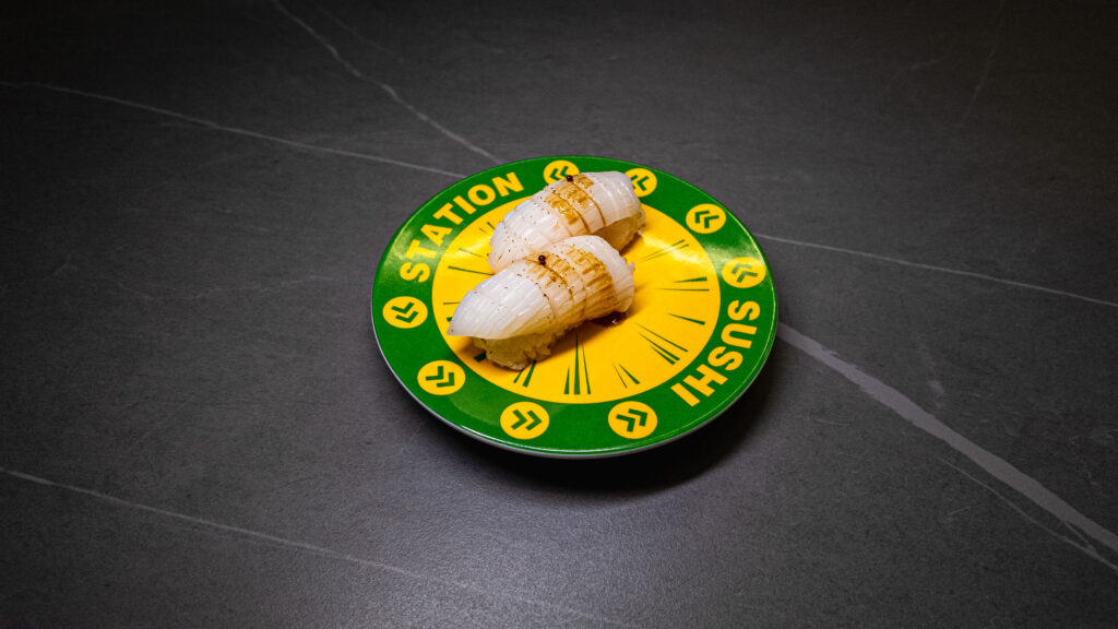 Sushi nigiri on green and yellow clock-themed plate at restaurant on dark tile surface