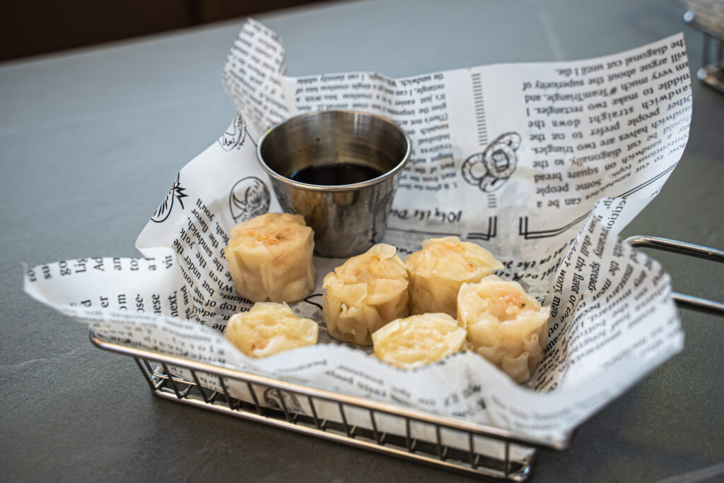 Steamed dumplings served in wire basket lined with newspaper alongside small metal dipping sauce cup at restaurant