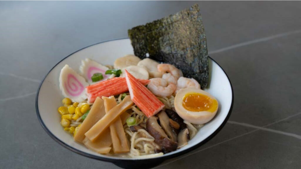 Bowl of ramen with shrimp, soft-boiled egg, nori, crab sticks, corn, mushrooms, and fish cakes at CROWDFAVORITES restaurant
