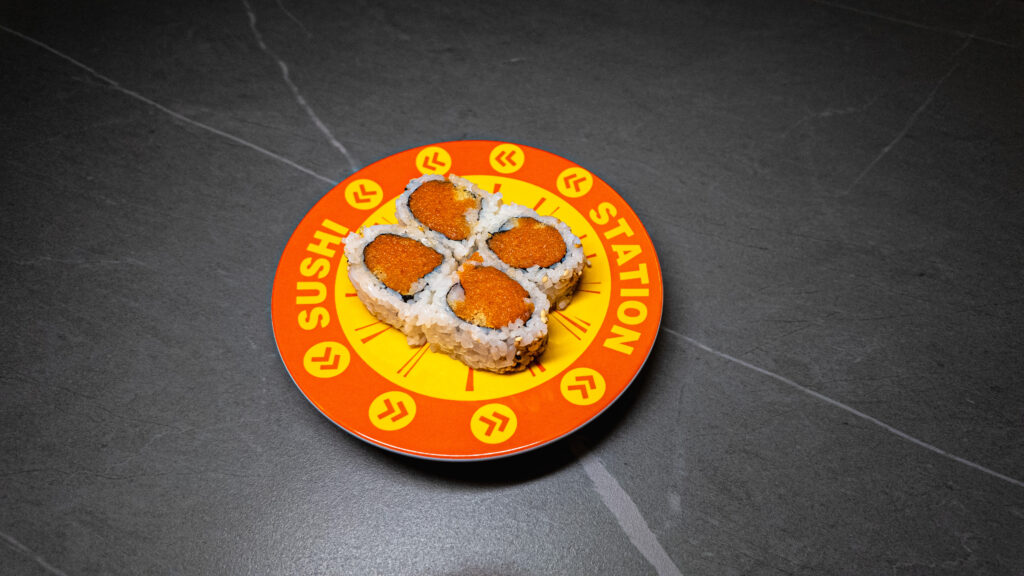 Four sushi rolls with orange topping on vibrant orange “Sushi Station” plate, served at CROWDFAVORITES restaurant.