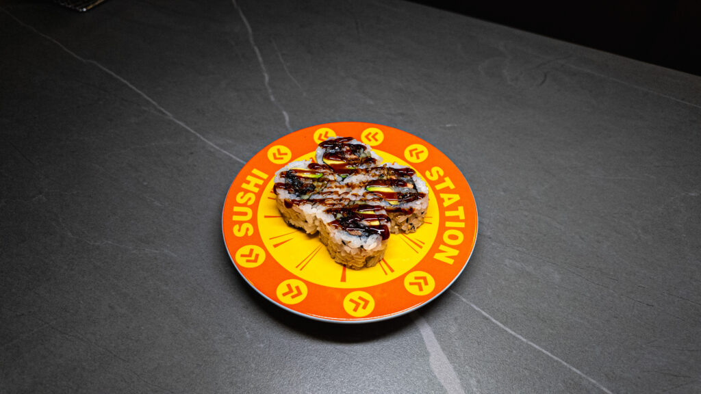 Sushi Station plate with four sushi rolls drizzled in sauce, served on vibrant orange and yellow dish at CROWDFAVORITES re...