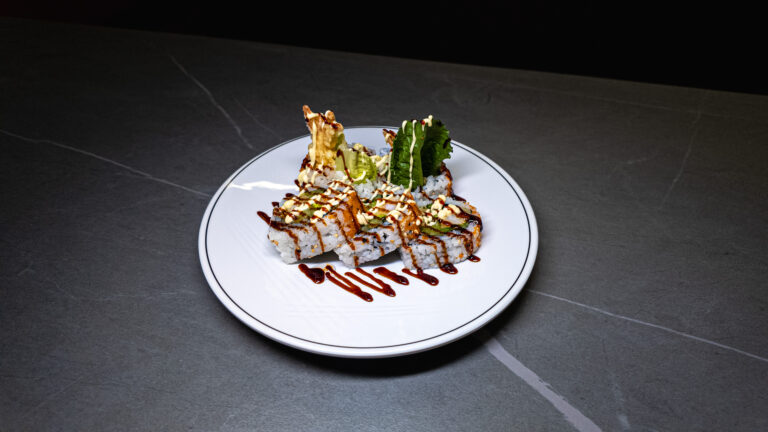 Gourmet sushi roll with tempura, drizzled sauce, and fresh greens on white plate — CROWDFAVORITES restaurant specialty