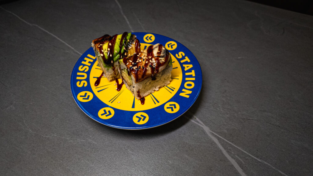 Sushi rolls with avocado, eel, and sesame seeds on a blue "Sushi Station" plate, drizzled with sauce, at CROWDFAVORITES re...