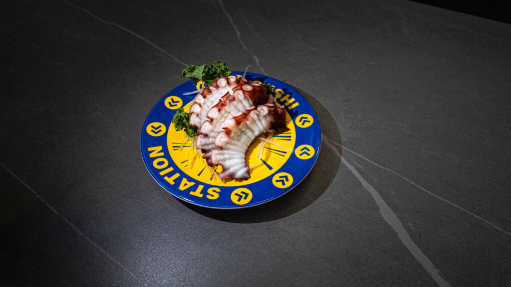 Sliced octopus sashimi on blue and yellow “STATION” plate, garnished with greens, served at CROWDFAVORITES restaurant.