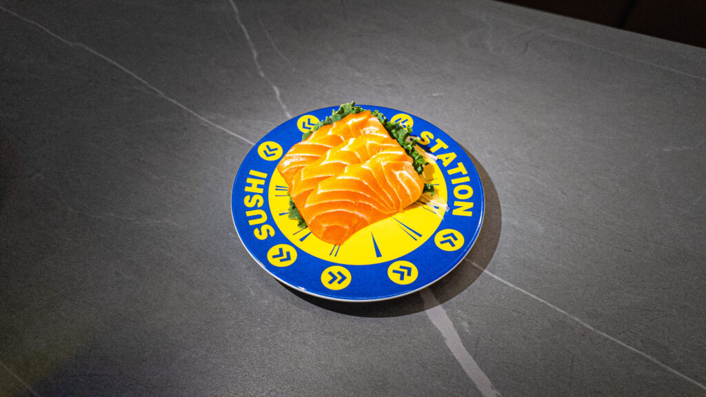 Fresh salmon sashimi on blue and yellow Sushi Station plate, served on dark table at CROWDFAVORITES restaurant.